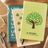 Tre notebook ecologici con copertina in carta semi di diversi colori (verde scuro, beige, verde chiaro) con logo Reforest e icone naturali, appoggiati su un tavolo di legno accanto a forbici e spago.