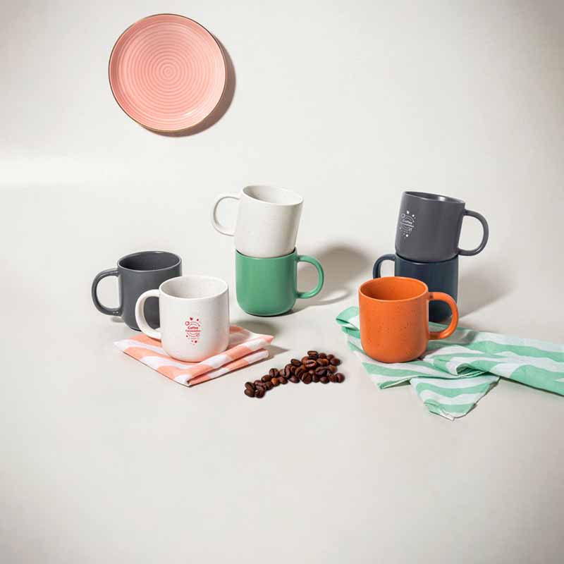 Composizione lifestyle di tazze in ceramica colorate impilate con chicchi di caffè e piattino decorativo.