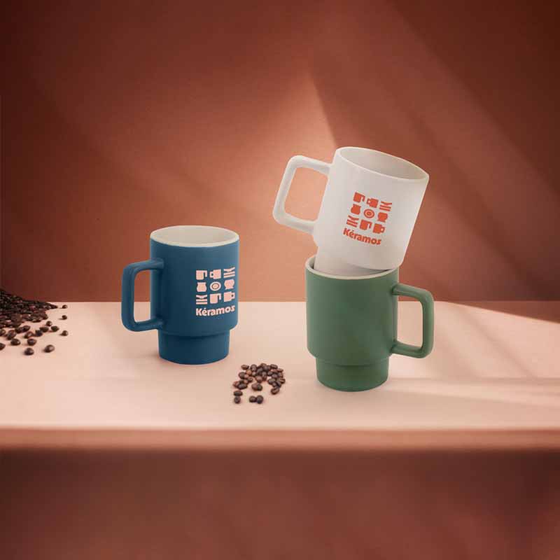 Ambientazione lifestyle con tazze Keramos blu, bianche e verdi su un tavolo con chicchi di caffè, tazze di design impilabili.